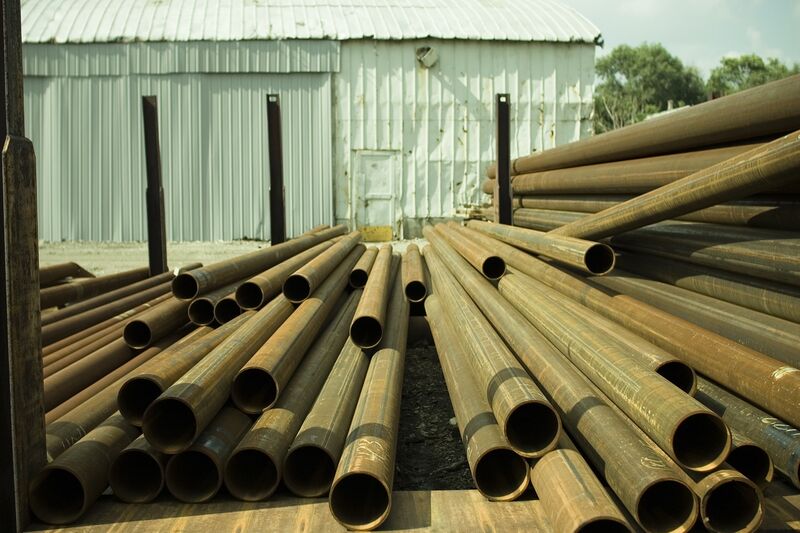 Rows of rusty pipes lying in a yard in front of a metal shed, with a pile of larger diameter pipes on the right